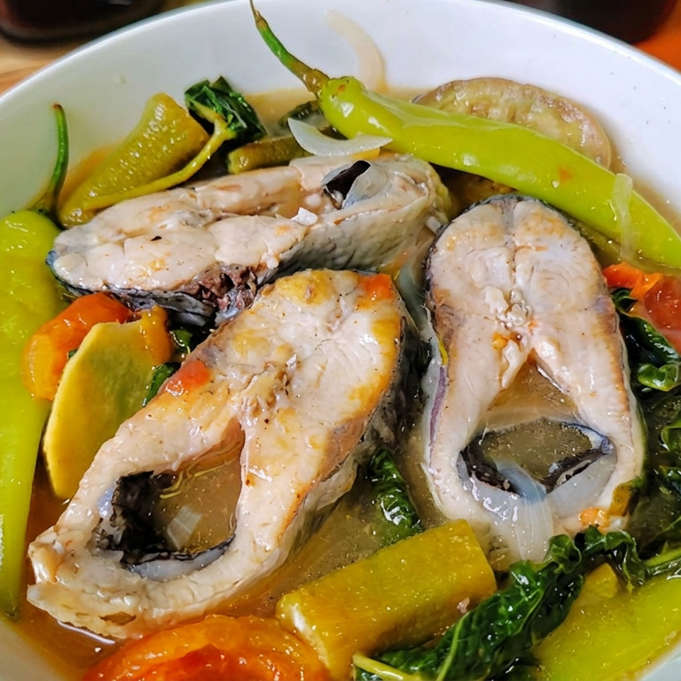 Bowl of fish steaks in savory broth with green peppers, tomatoes, onions, and leafy greens.